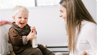 A smiling toddler in brown overalls holds a bottle while sitting on a chair, laughing with a woman who is facing him and smiling. They appear to be enjoying a happy moment together indoors.