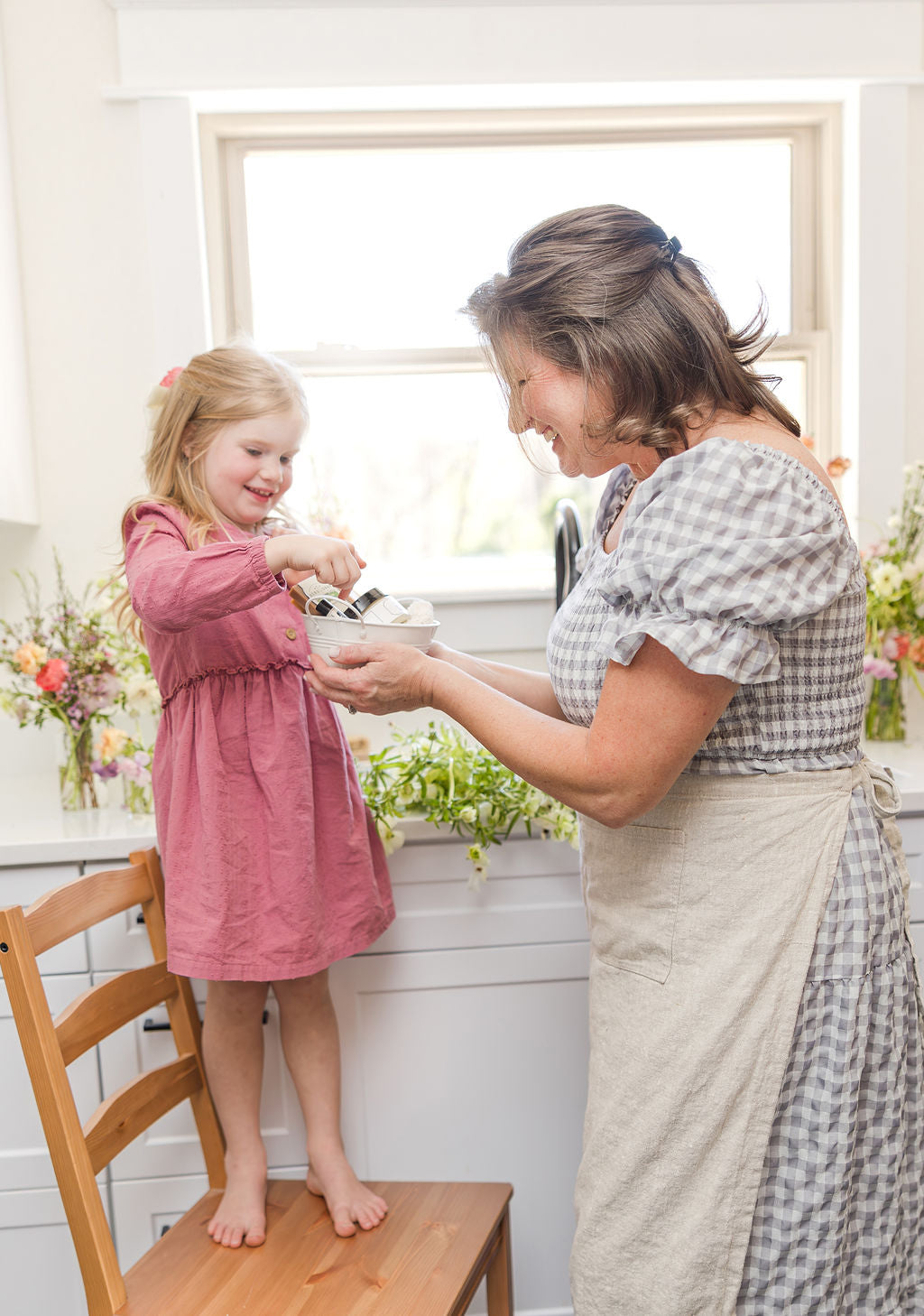 A young girl in a pink dress stands on a wooden chair, cracking eggs into a bowl held by a smiling woman in a checkered dress and apron in a bright kitchen with flowers on the windowsill.