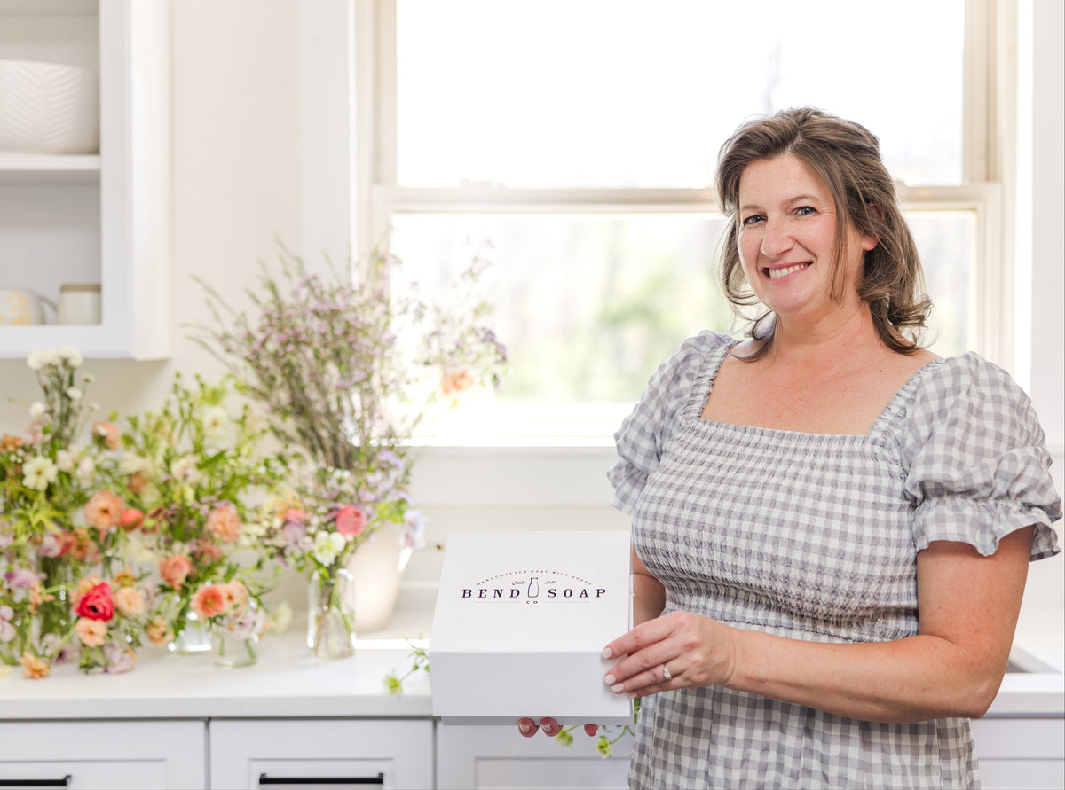 A smiling woman in a gray gingham dress stands in a bright kitchen, holding a white box labeled "Bend Soap." Behind her, a counter is decorated with colorful flowers in vases.