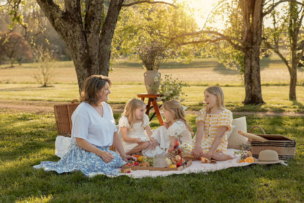 A woman and three young girls sit on a blanket having a picnic under trees in a sunlit park, surrounded by baskets and snacks. They are smiling and enjoying each other's company on a warm day.