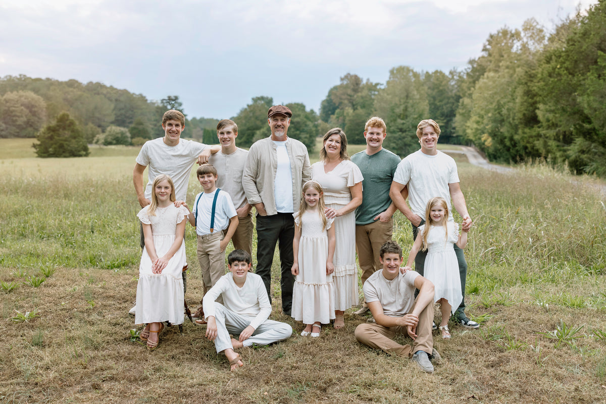 A family group of twelve—adults, teens, and children—pose outdoors in a grassy field with trees and a winding road in the background. They wear coordinated light-colored casual outfits and smile at the camera.