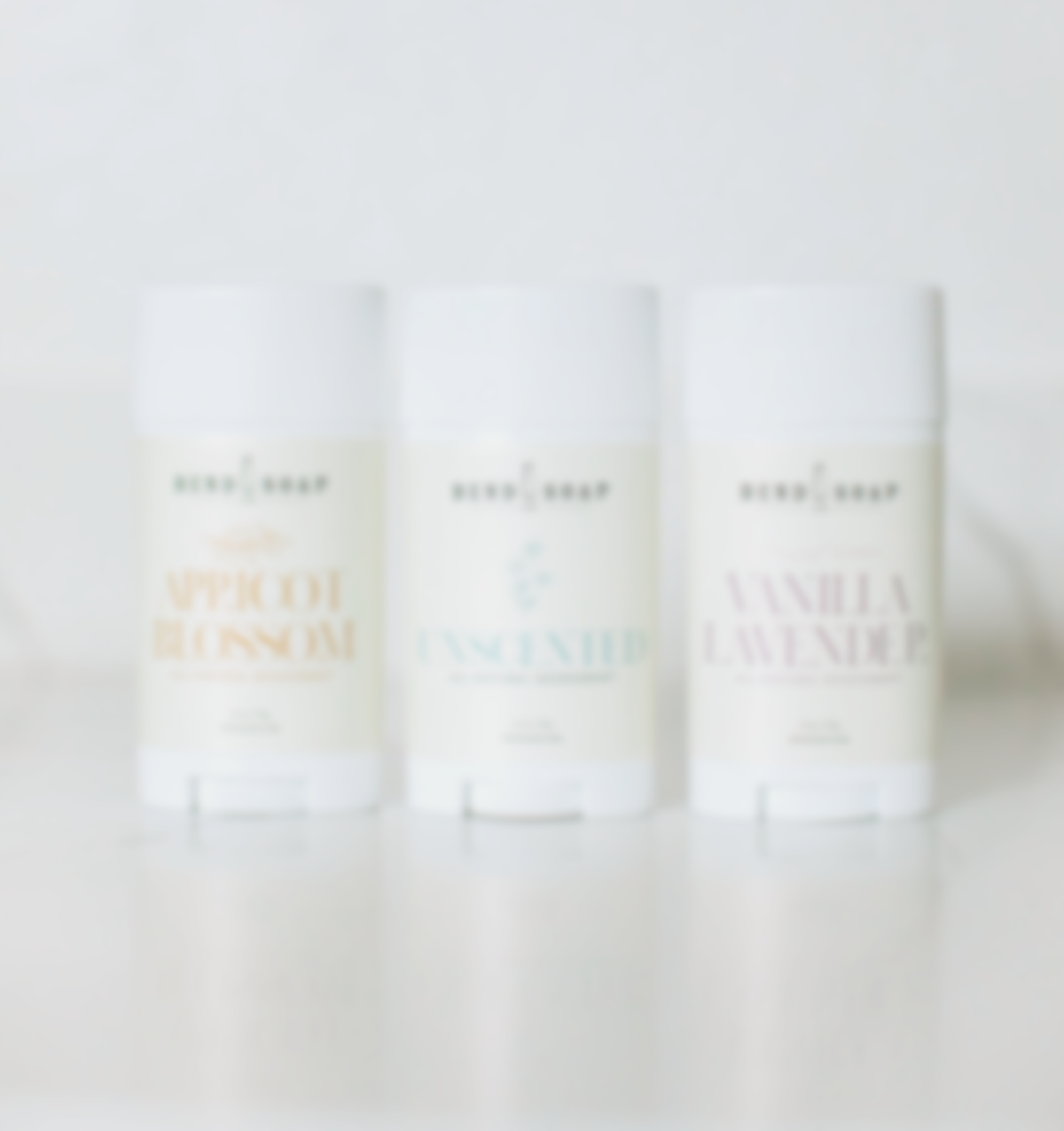 Three white deodorant sticks labeled "Apricot Blossom," "Unscented," and "Vanilla Lavender" are displayed side by side on a white surface. The image is softly focused and bright.