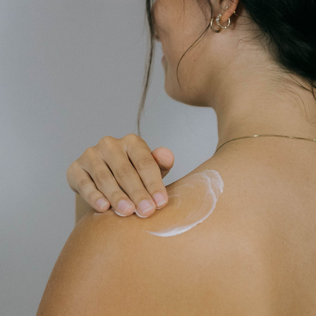 A person with shoulder-length dark hair applies white lotion to their bare shoulder. The image is taken from behind, showing their hand spreading the lotion and a simple gold necklace around their neck.