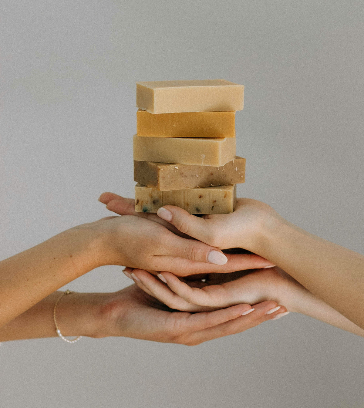 Several hands gently hold a stack of natural, handmade soap bars in various earthy colors against a plain, light background.
