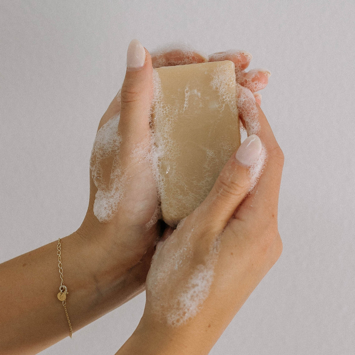 Light pink manicured hands hold a Peppermint Goat Milk Soap Scrap (1 lb.), lathered with white foam against a plain background. A delicate gold bracelet adds elegance, highlighting this artisanal cleanser.