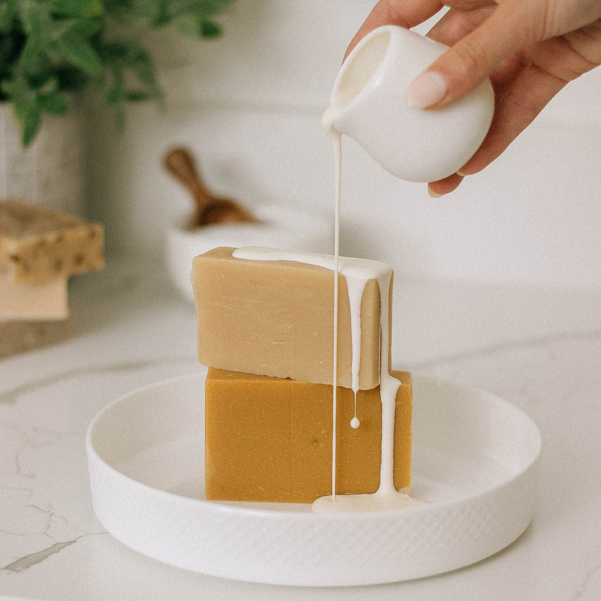 A hand pours a creamy white liquid over two stacked Lavender Blossom Goat Milk Soap Scraps (1 lb.) on a white plate, with more handcrafted soap and greenery blurred in the background.