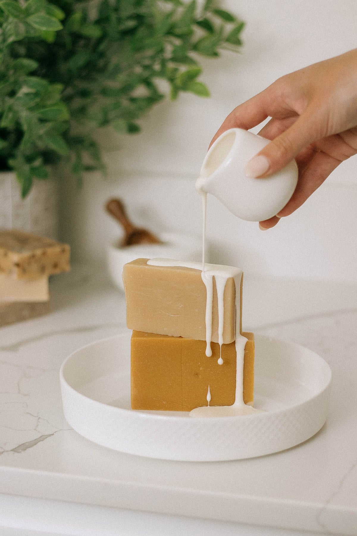 A hand pours white liquid from a small pitcher onto two stacked blocks of soap on a white plate, with a green plant blurred in the background.