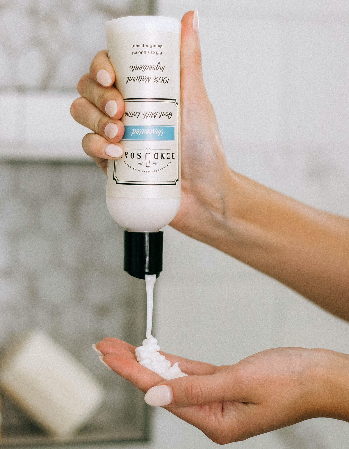 A person dispenses white lotion from a BEND SOAP CO. bottle onto their palm, with a bar of soap blurred in the background.