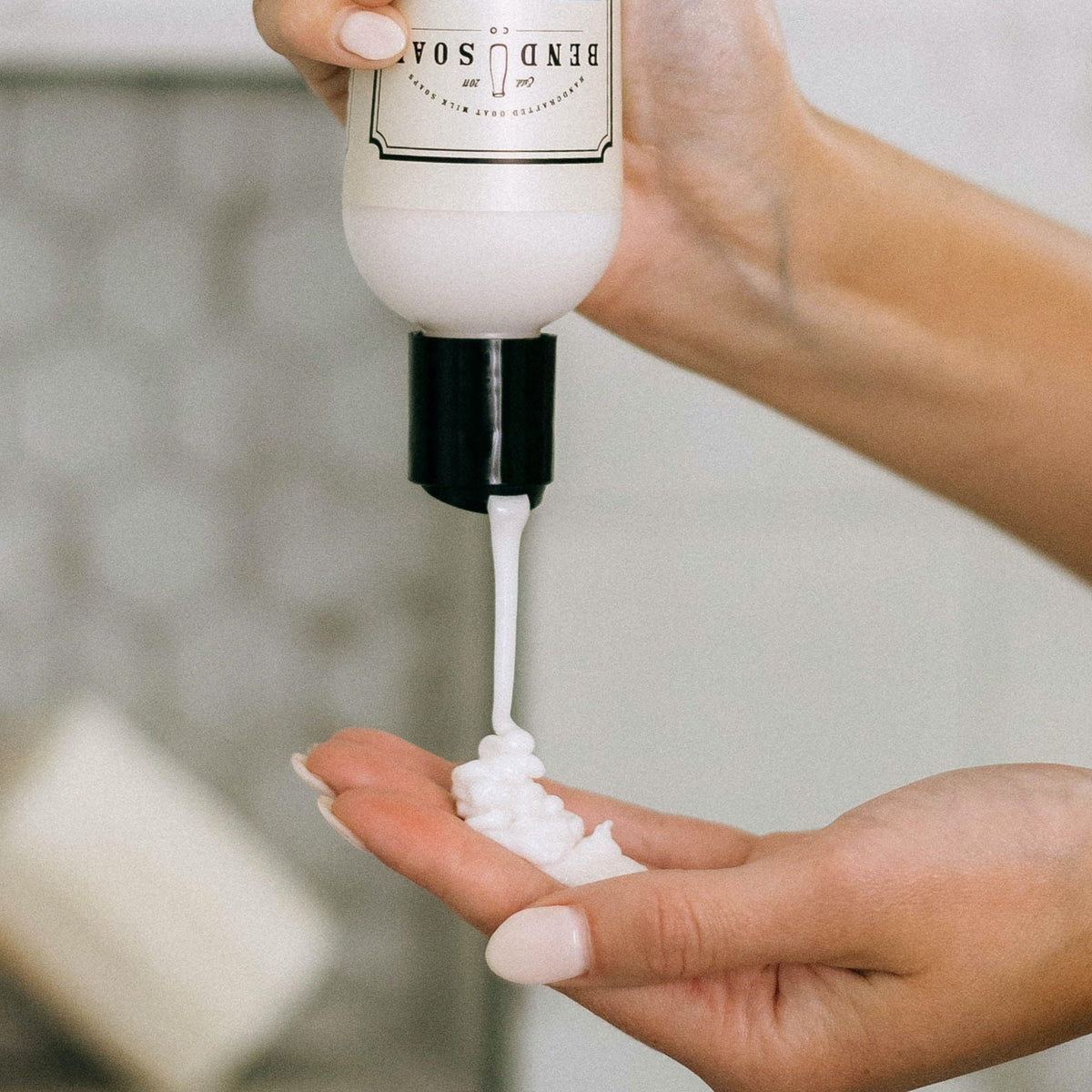 A person squeezes white lotion or soap from a bottle labeled "BEND SOAP" onto their palm, with both hands visible against a blurred background.