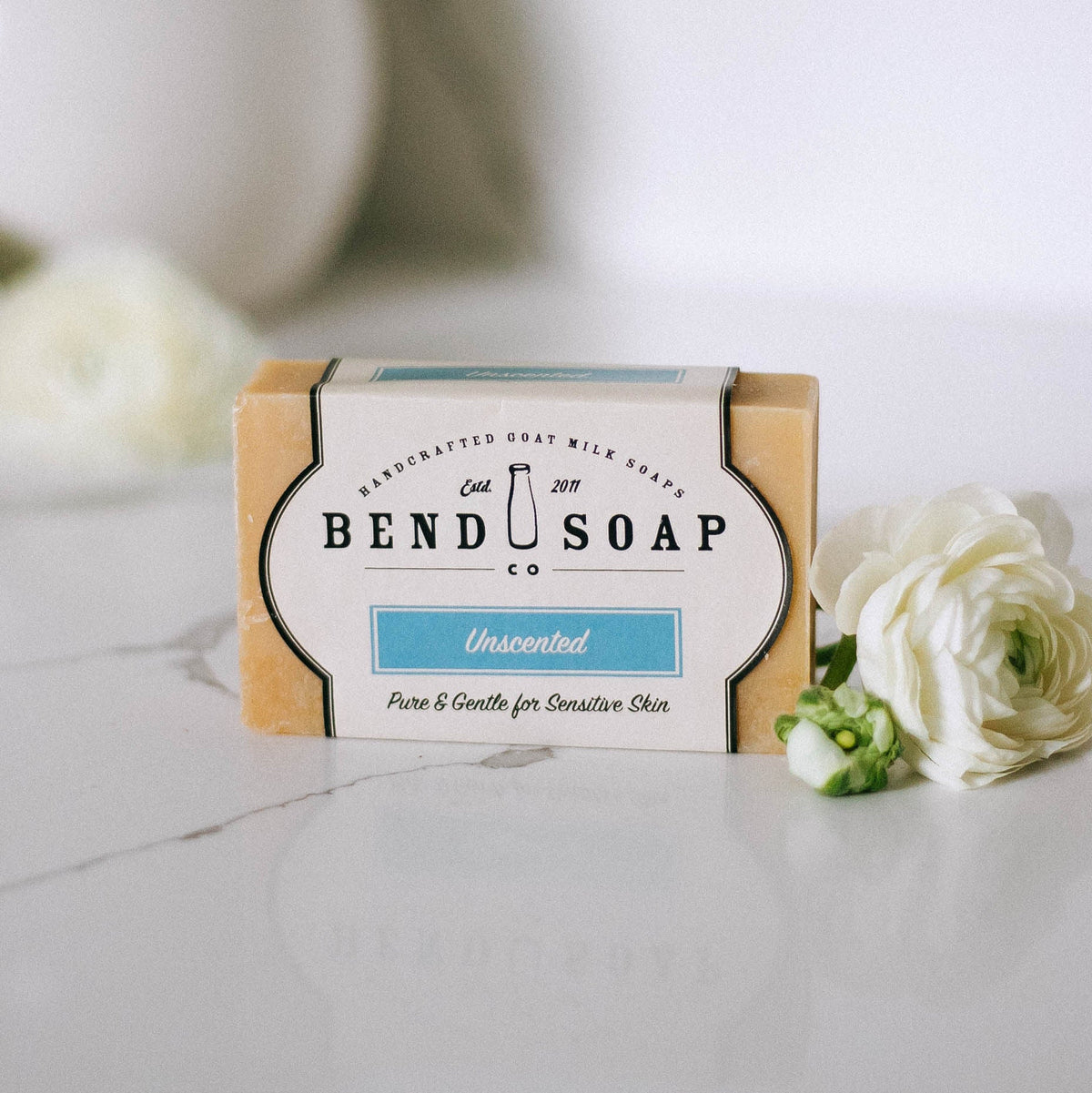 A bar of Bend Soap Co. unscented goat milk soap sits on a white surface, with white flowers nearby. The label reads “Pure & Gentle for Sensitive Skin.”.