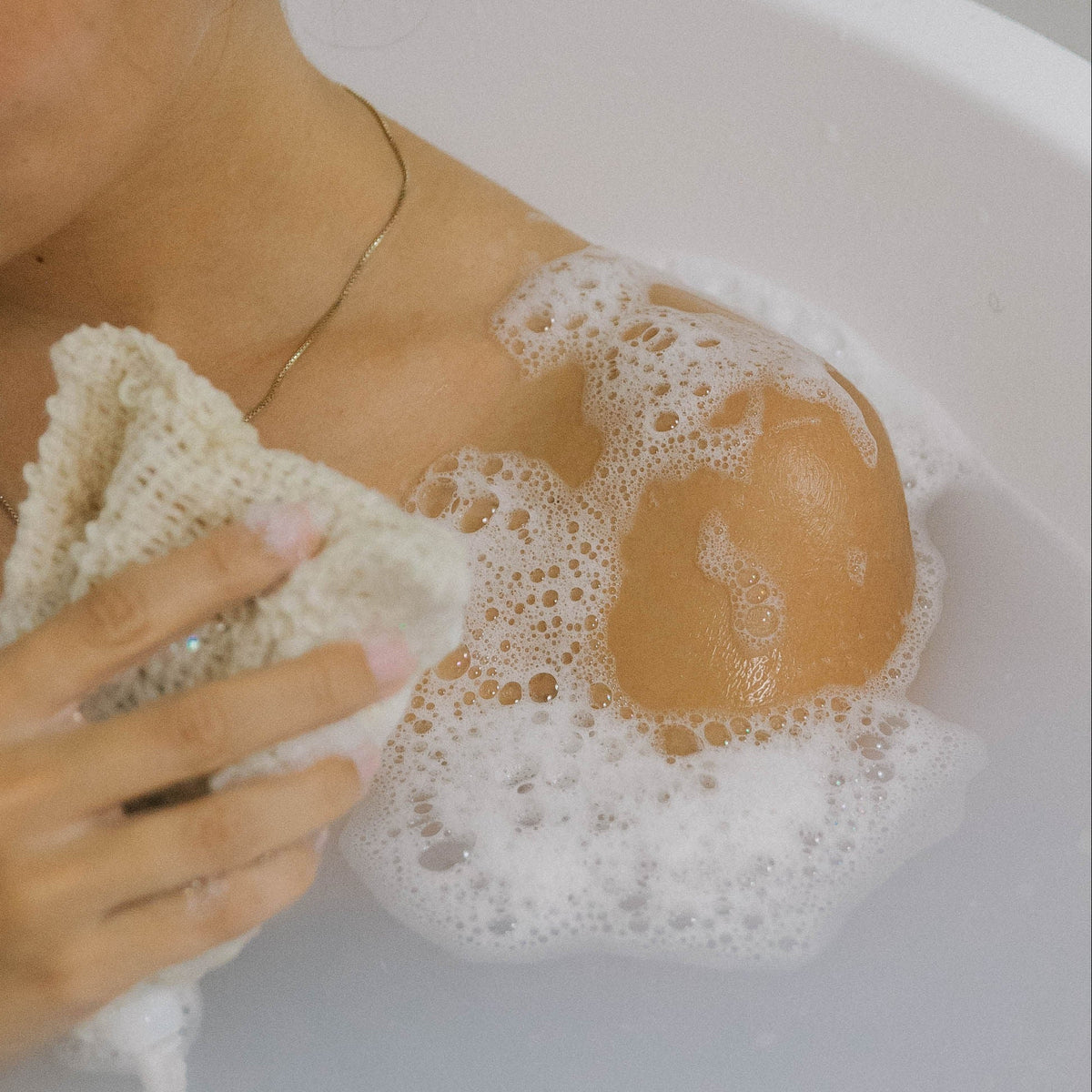 A person in a bathtub holds a textured washcloth, gently scrubbing their shoulder, which is covered in soap suds and bubbles. Only part of their shoulder, neck, and hand are visible.