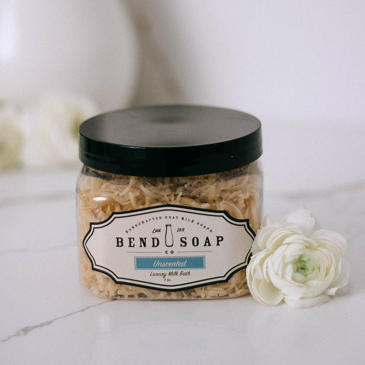 A clear jar of Bend Soap Company Unscented Luxury Milk Bath sits on a white surface next to a white flower. The jar has a black lid and shredded bath product inside.