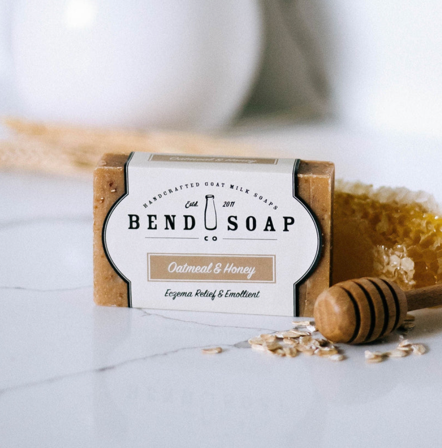 A 12 Pack of Imperfect Bars - Oatmeal & Honey Goat Milk Soap is shown with honey dipper, honeycomb, oats, and wheat. The label highlights its gentle formula for eczema relief and sensitive skin.