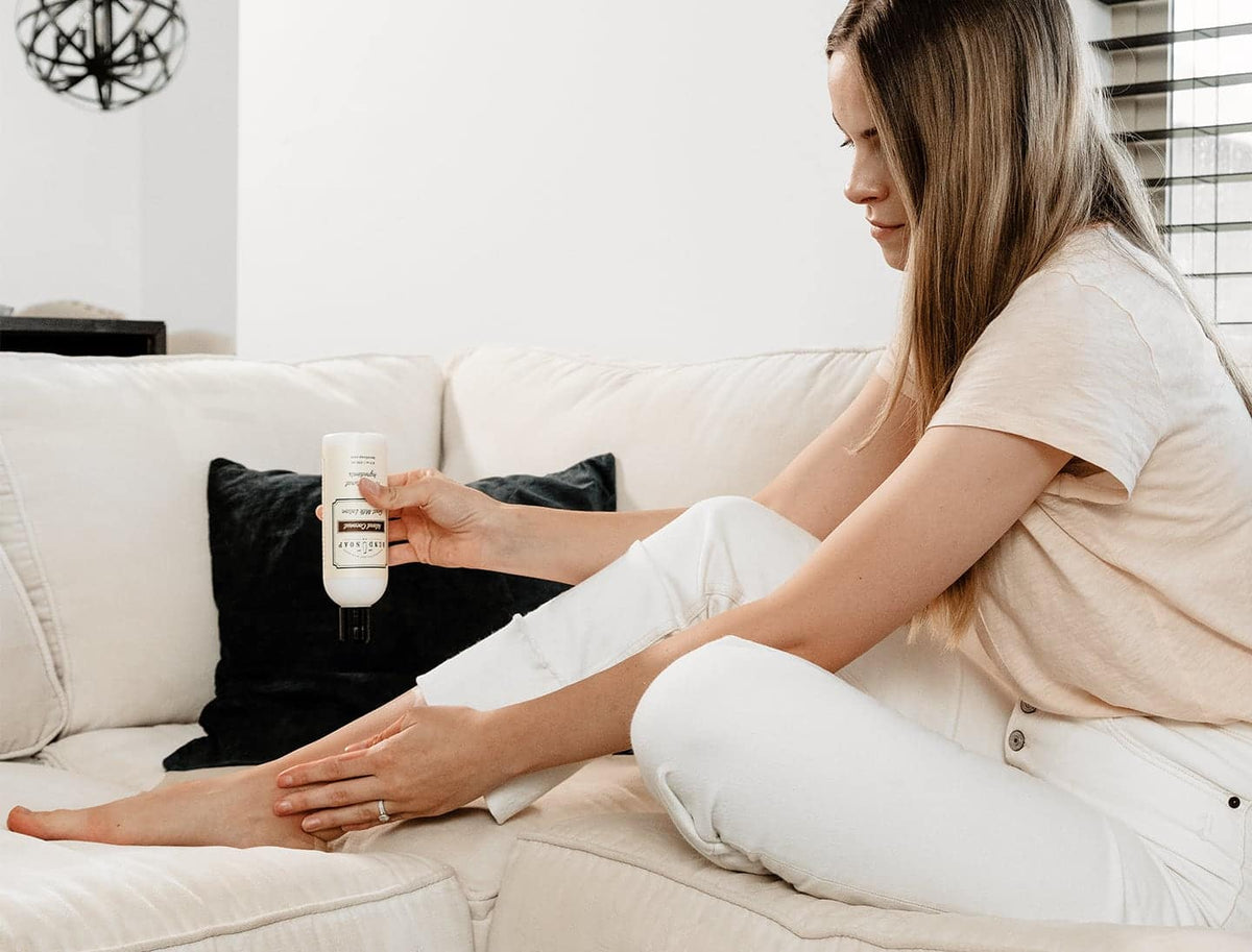 A woman sitting on a white couch applies lotion from a small bottle to her lower leg. She is wearing a light-colored shirt and white pants, and there is a black cushion behind her.