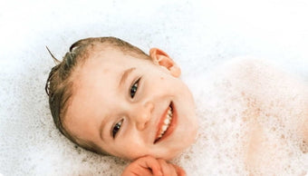 A young child with wet hair smiles while lying in a bubble bath, surrounded by white suds.