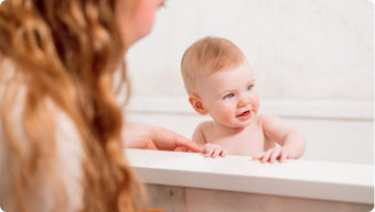 A baby with light hair sits in a bathtub, smiling and looking to the side, while an adult with long wavy hair is partially visible beside the tub, gently holding the baby's arm.