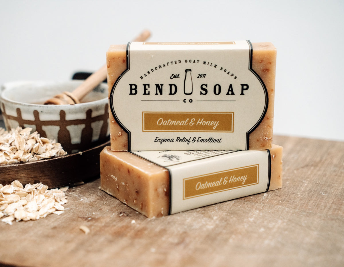 Two bars of Bend Soap Co. oatmeal and honey goat milk soap sit on a wooden surface near a bowl of oats and a honey dipper, with some loose oats scattered nearby.