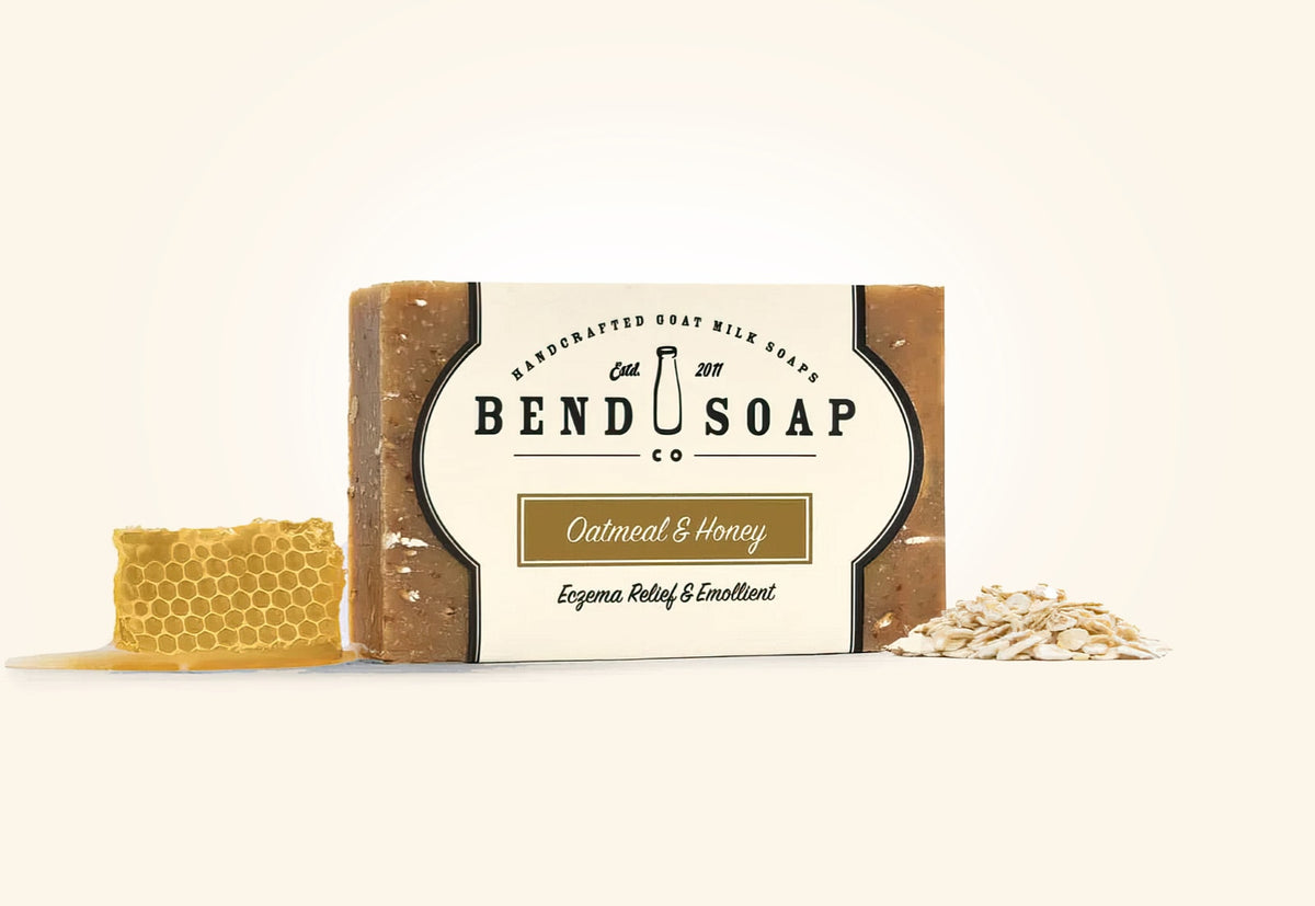 A bar of Bend Soap Co. Oatmeal & Honey handcrafted goat milk soap is shown with a honeycomb piece and a small pile of oats beside it. The soap label mentions relief for eczema and being emollient.