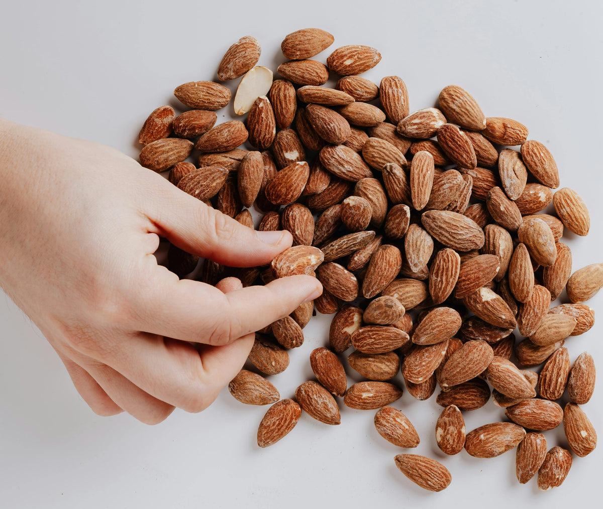 A hand picking up a raw almond from a pile of unshelled almonds on a white surface.