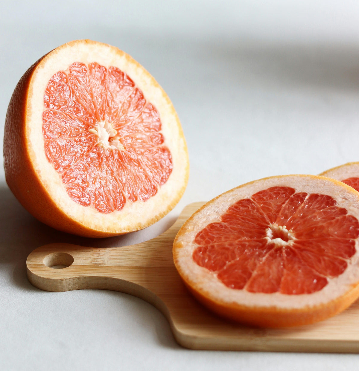 A halved grapefruit and two grapefruit slices rest on a small wooden cutting board against a white background. The fruit’s pinkish-orange flesh and thick rind are clearly visible.