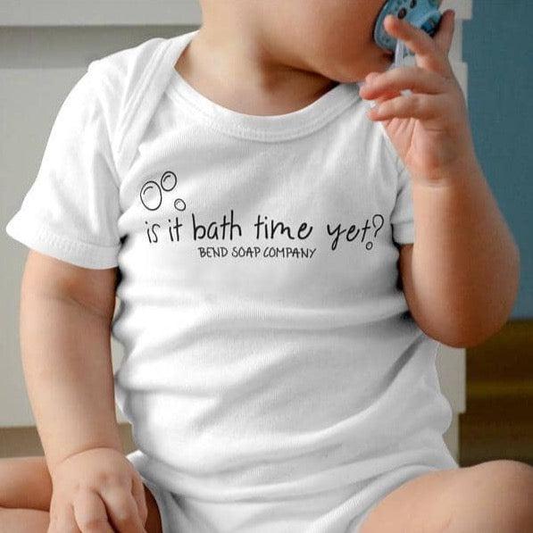 A baby wearing a 100% cotton Baby Onesie with "is it bath time yet? BEND SOAP COMPANY" on the front, holding a pacifier. Features easy snap fasteners for quick changes.